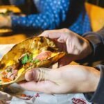 Close-up of a person holding a fresh Mexican taco filled with meat, vegetables, and herbs at a restaurant table.