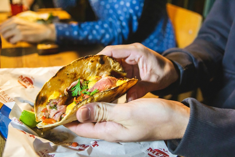 Close-up of a person holding a fresh Mexican taco filled with meat, vegetables, and herbs at a restaurant table.