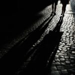 Silhouettes of two people walking at night on a dimly lit cobblestone street, casting long shadows under streetlight.