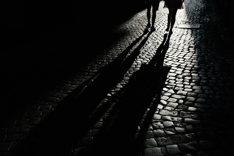Silhouettes of two people walking at night on a dimly lit cobblestone street, casting long shadows under streetlight.
