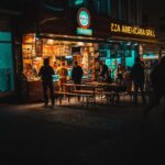 Late-night street food city scene showing people ordering from a neon-lit food stall on a busy urban street at night.
