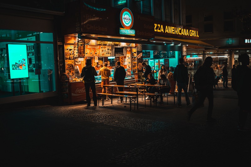 Late-night street food city scene showing people ordering from a neon-lit food stall on a busy urban street at night.